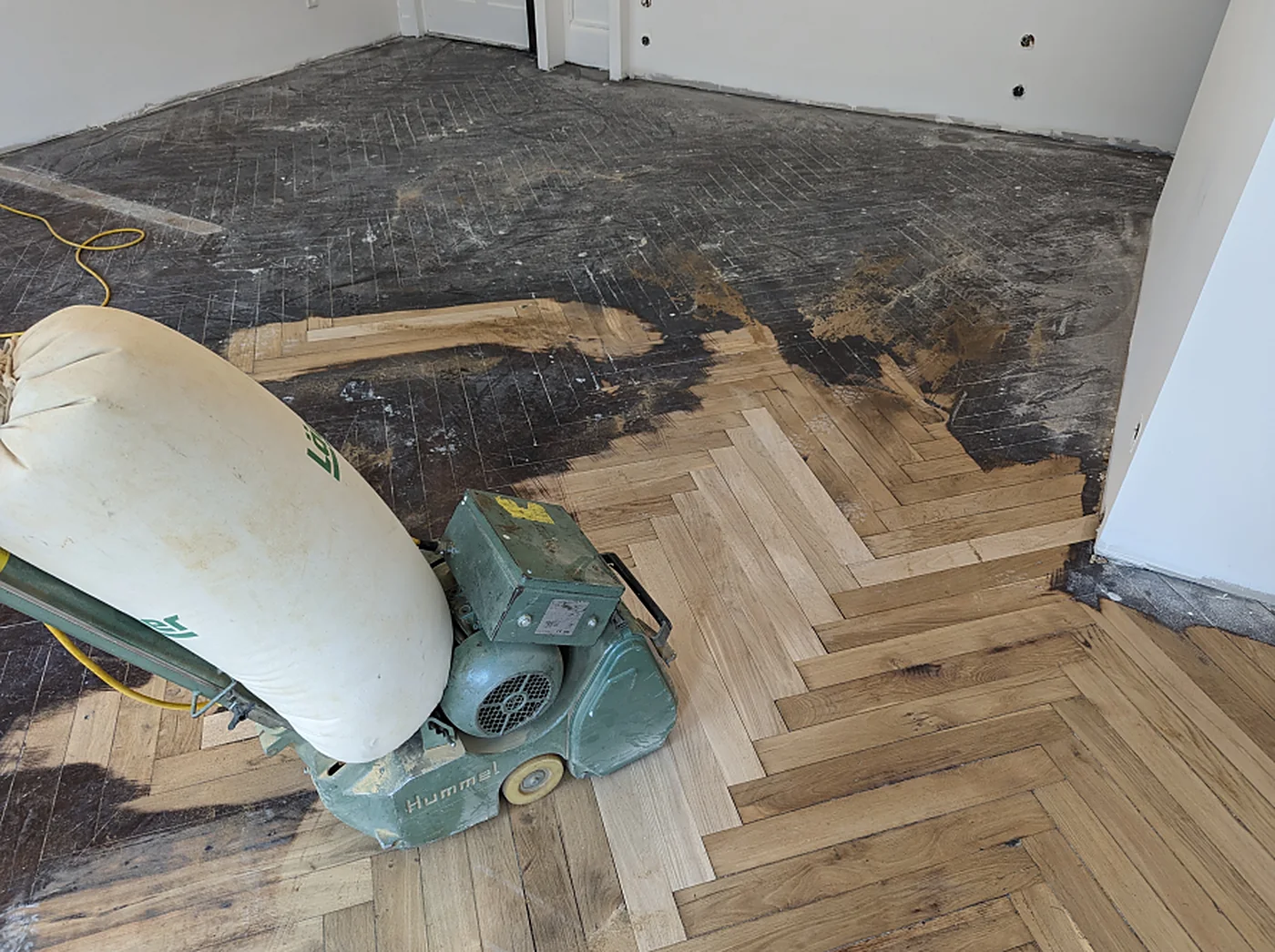 Ponceuse à bande avec son sac d’aspiration sur un parquet en chevrons à moitié décapé.