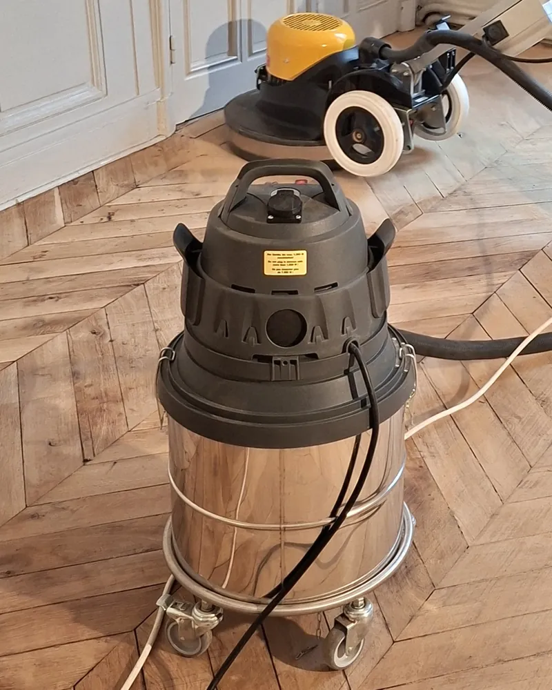 Aspirateur industriel en inox raccordé à la ponceuse, sur un parquet en chevrons.