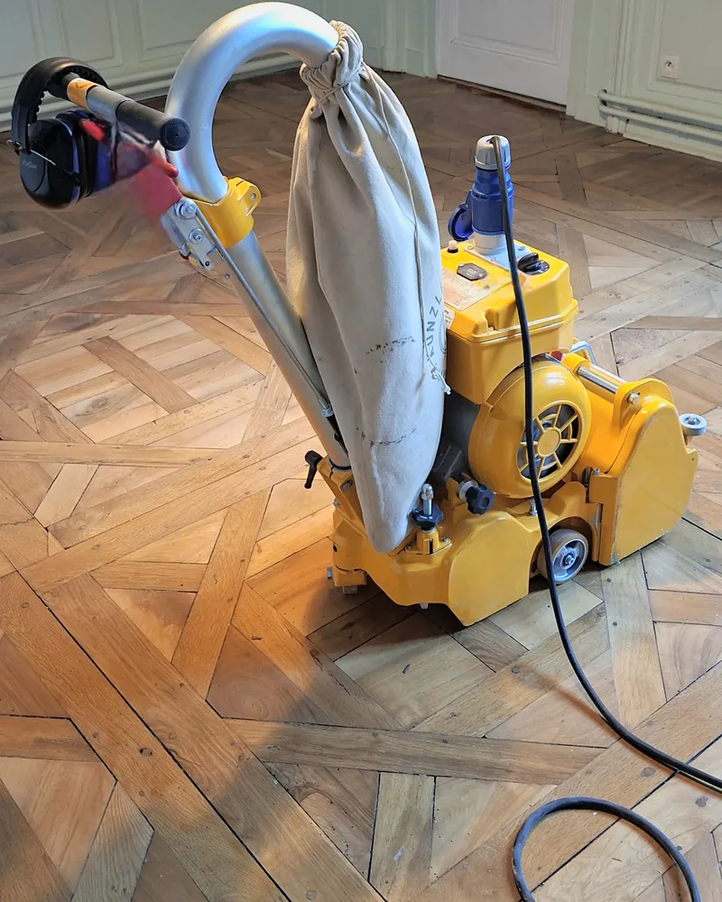 Ponceuse à bande jaune avec son sac d’aspiration sur un parquet à compartiments.